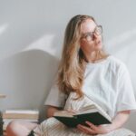 Frau sitzt lesend auf dem Boden mit Bücherstapeln neben ihr